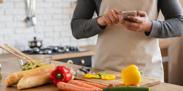 Mann steht in der Küche vor Lebensmitteln und schaut in sein Smartphone