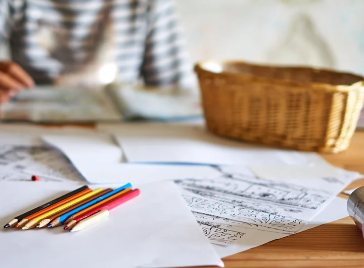 Person sitzt am Tisch und malt, auf dem Tisch liegen Stifte und Papier