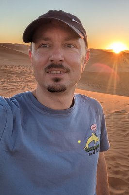 Profilfoto von Robert, Mann mit Bart und Käppi steht in der Wüste bei Sonnenuntergang und lächelt in die Kamera