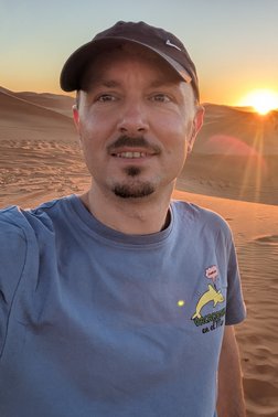 Profilfoto von Robert, Mann mit Bart und Käppi steht in der Wüste bei Sonnenuntergang und lächelt in die Kamera