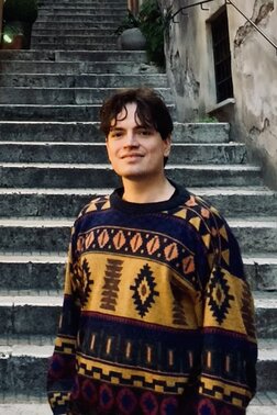 Junger Mann mit buntem Pullover lächelt vor alter Steintreppe in schmaler Gasse